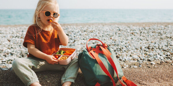 Child girl with lunchbox eating vegetables outdoor travel vacation healthy lifestyle vegan food picnic on beach hungry kid with lunch box snacks and backpack SA_Reiseapotheke_fuer_Kinder